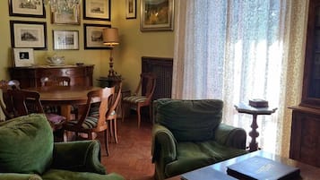 Een klassieke zithoek met groen gestoffeerde meubels, een kroonluchter, ingelijste foto's aan de wanden, en een eethoek met een houten tafel en stoelen.
