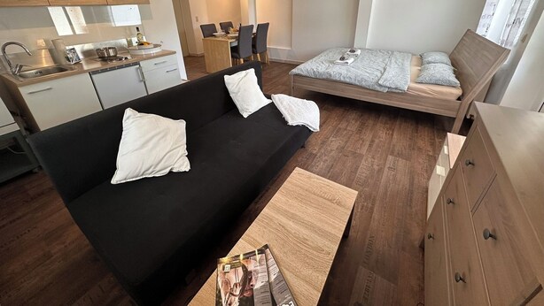 A compact living space with a kitchenette, a black sofa with white cushions, a wooden bed, and a wooden dresser.