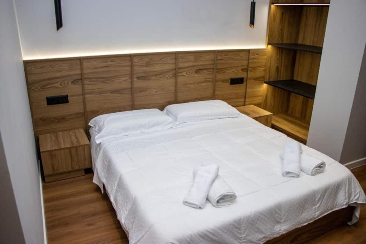 Una cama bien hecha con sábanas blancas y toallas enrolladas, un cabecero de madera y un mueble estante empotrado.