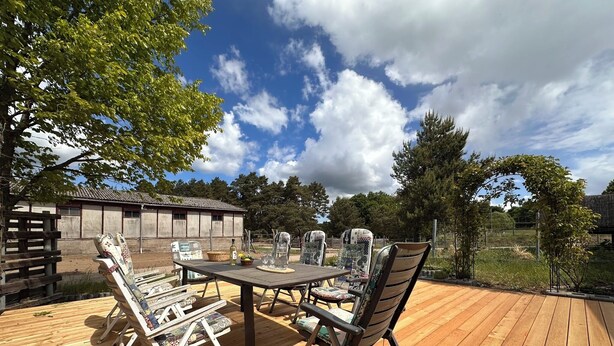 An outdoor wooden deck with a table and chairs, surrounded by trees and a building in the background. - Modern Holiday room with Garden and Horse Riding near Warnow (Warnow)
