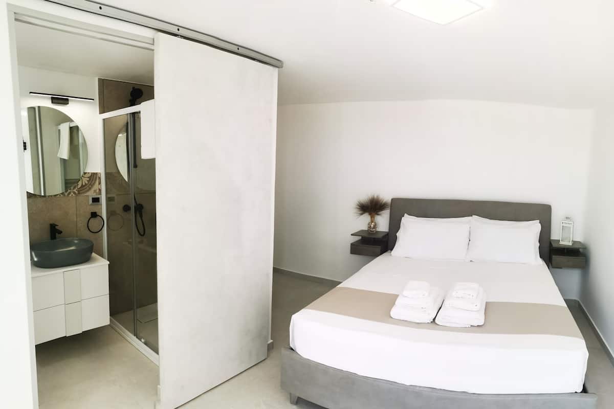 A modern bedroom with a bed, bedside tables, a bathroom with a sink and mirror, and a sliding door to a shower area.