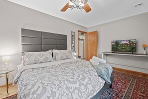 Une chambre avec un grand lit, un ventilateur de plafond, un téléviseur posé sur une étagère et une porte donnant sur une autre pièce.