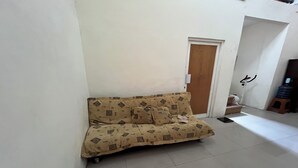 A room with a beige sofa against a white wall, a wooden door, and a small stool. - Spot on Homestay Griya Sunarti (Depok)
