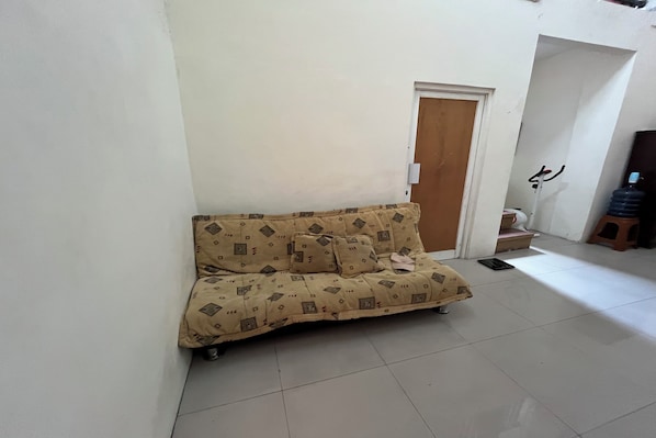 Una stanza con un divano color beige affacciato su una parete bianca, una porta in legno e uno sgabello.