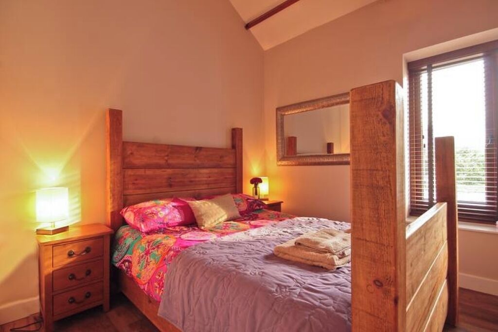 Un dormitorio con cabecera de madera, cama con ropa de cama colorida, mesita de noche con lámpara y ventana con persianas.