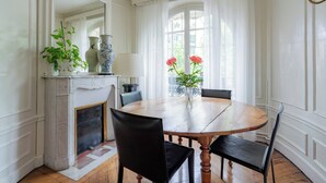 A dining room with a round wooden table, black chairs, a vase with flowers, a fireplace, and a large window with curtains.