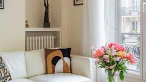 A living room with a white sofa, a glass coffee table, a colorful rug, a vase with flowers, and a window with a view of buildings.