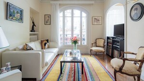 A living room with a white sofa, a glass coffee table, and a colorful rug. There are two armchairs and a TV stand with a bookshelf.