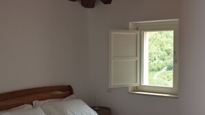 A bedroom with a wooden ceiling, a bed with white linens, a window with a view of greenery, and a bedside table. - Luxurious retreat for your well-being (Valle dell'Angelo)