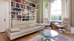 Un salon avec un canapé beige, une table basse en verre, une bibliothèque, deux fauteuils blancs et une grande fenêtre avec des rideaux.