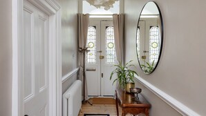 Un couloir avec une porte blanche, un radiateur, un banc en bois sur lequel se trouvent un bol et une plante, et un miroir accroché au mur.