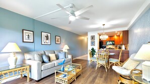 A modern living room with a sofa, coffee table, and chairs. A dining area with a table and chairs is visible in the background.