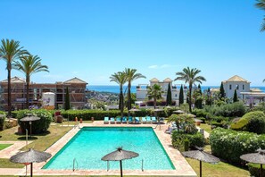 Zona de piscina con sillones de descanso y sombrillas, rodeada de palmeras y vista al mar.