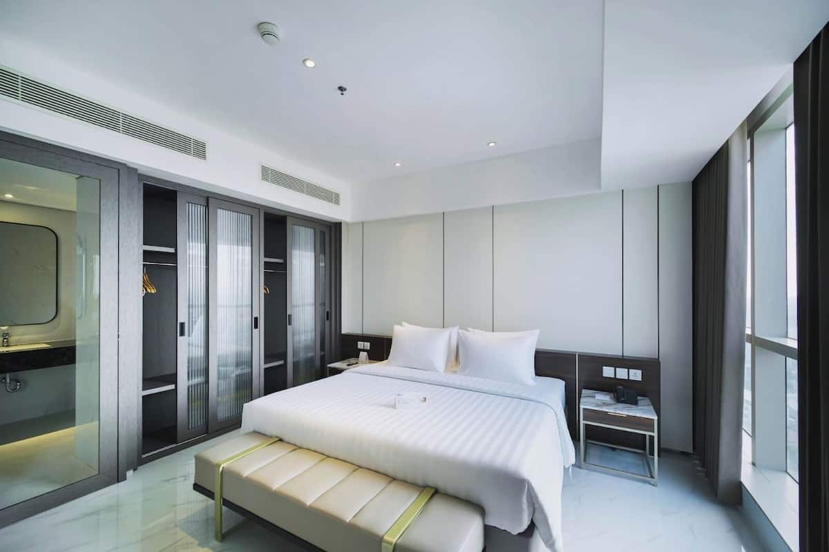 Una habitación de hotel moderna con una cama grande, una mesita de noche, un banco, un armario y un ventanal amplio.