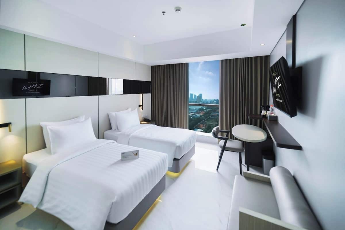 Habitación de hotel moderna con dos camas, un escritorio, una silla y un amplio ventanal con vistas a la ciudad.