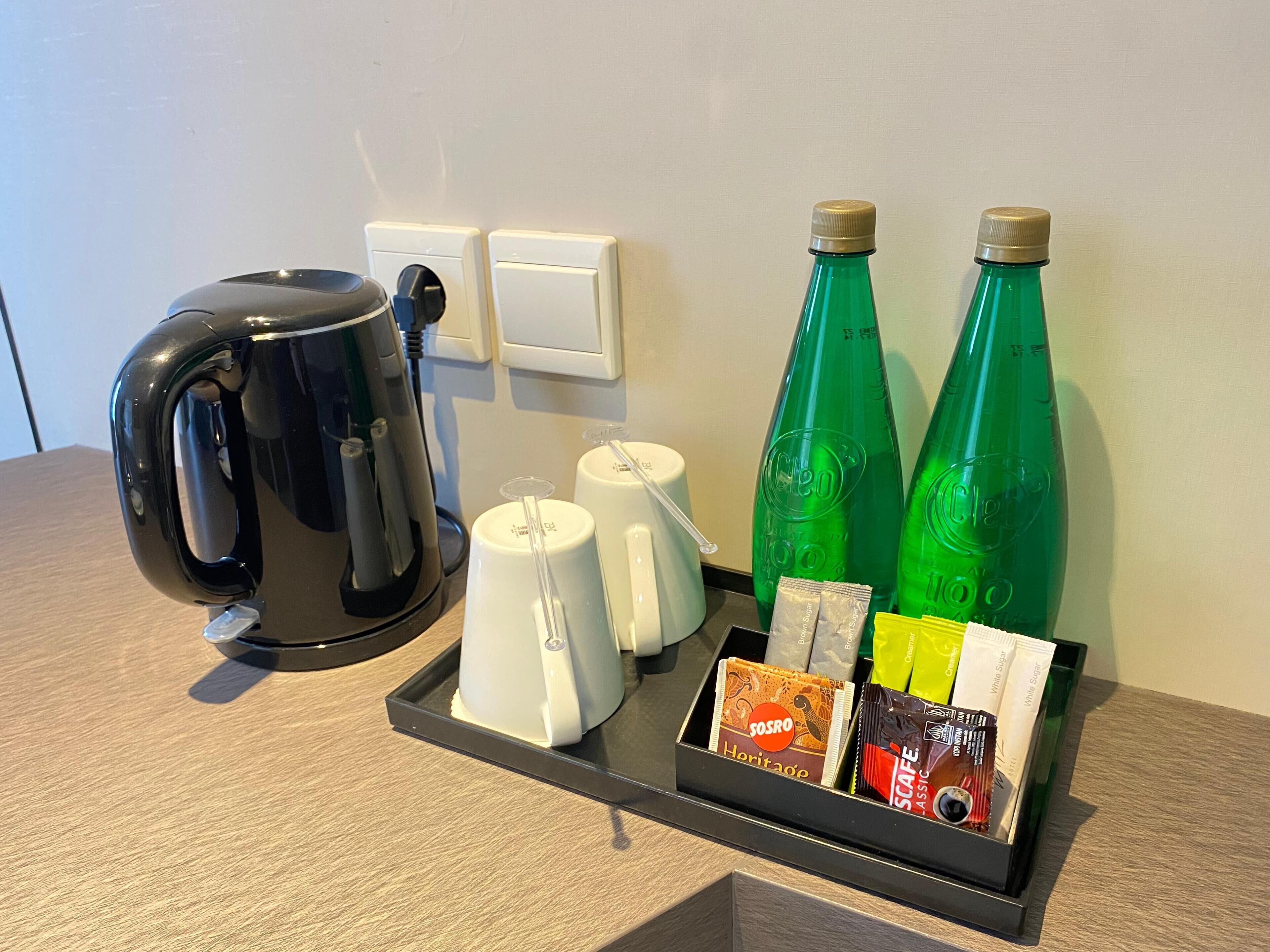 Een koffiezetapparaat, twee groene flessen, een witte mok en een dienblad met koffiepads op een hotelbalie.