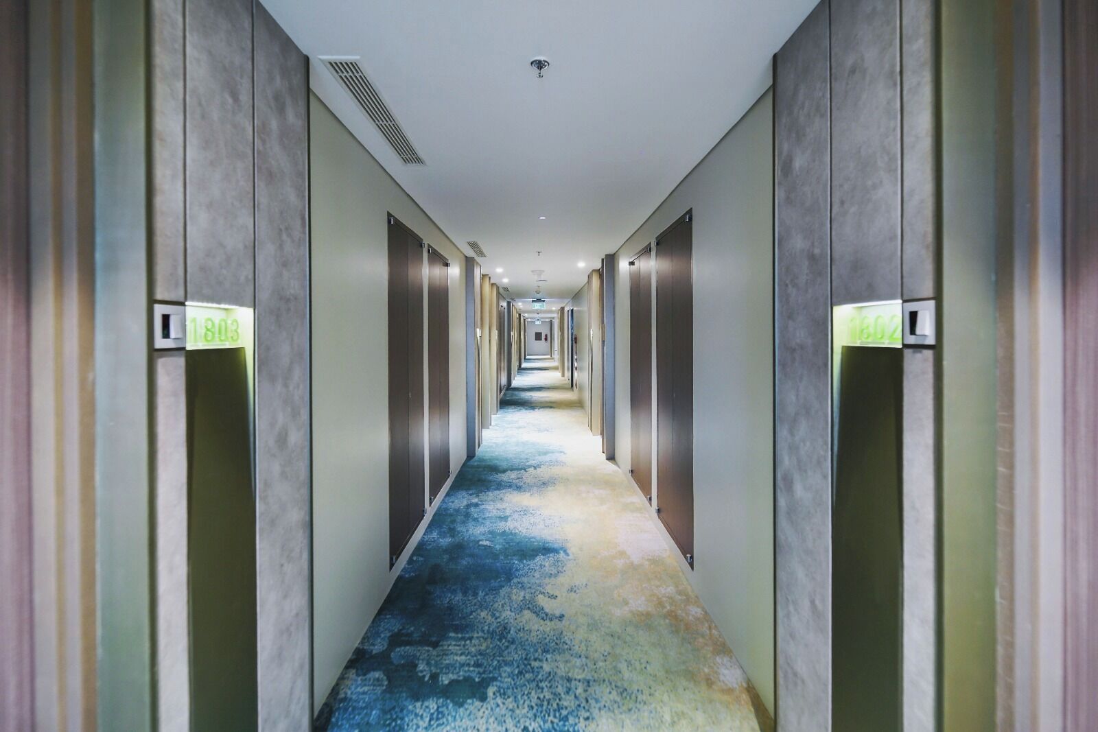 Un long couloir étroit d’hôtel, avec un tapis aux motifs bleus et jaunes, bordé de murs où se trouvent des portes d’ascenseur métalliques et des panneaux lumineux.