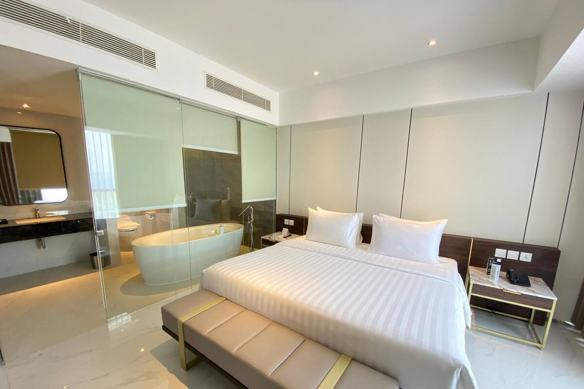 Habitación de hotel moderna con una cama grande, baño independiente con bañera, un banco y una mesita de noche con un teléfono y un libro.
