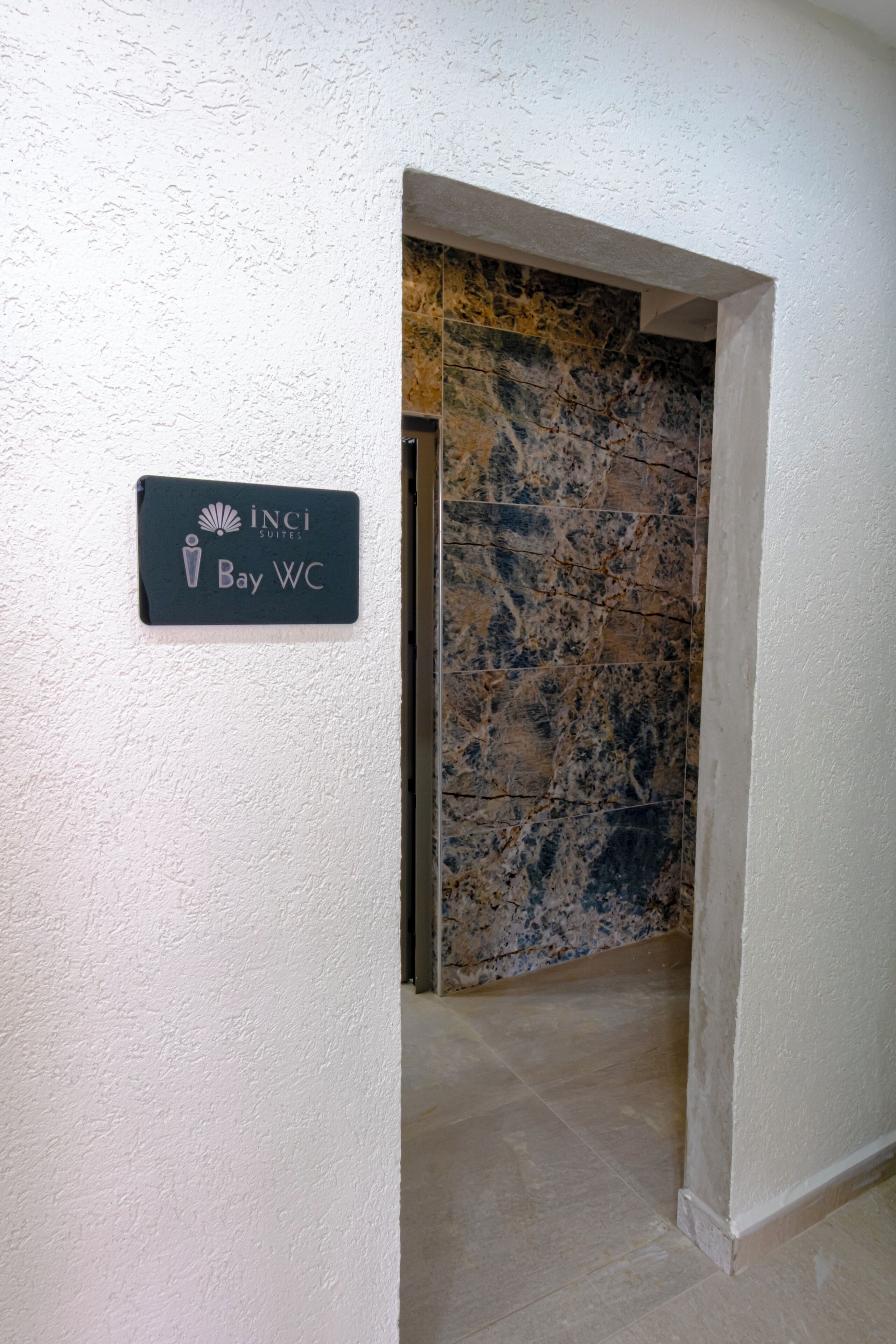A sign for 'Inci Suites Bay WC' in a hallway with a marble wall and a tiled floor.