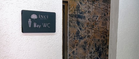 A sign for 'Inci Suites Bay WC' in a hallway with a marble wall and a tiled floor.