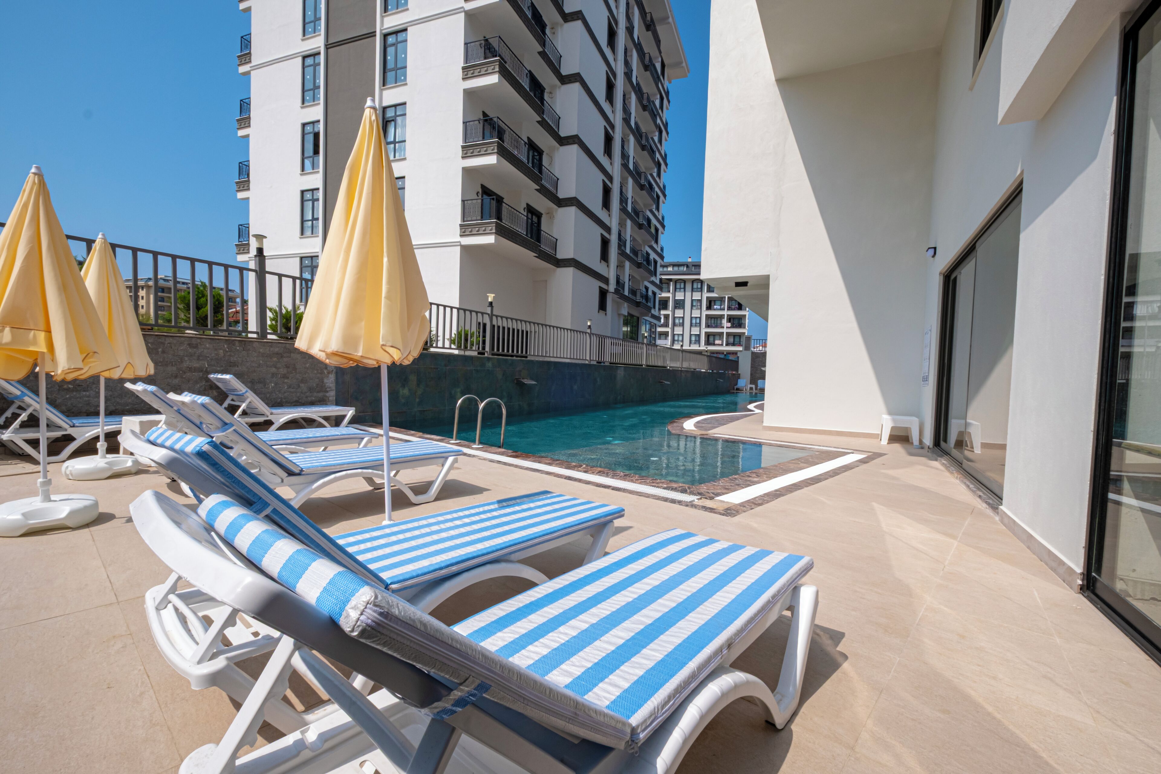 A poolside area with sun loungers and umbrellas, a pool, and a building with balconies.