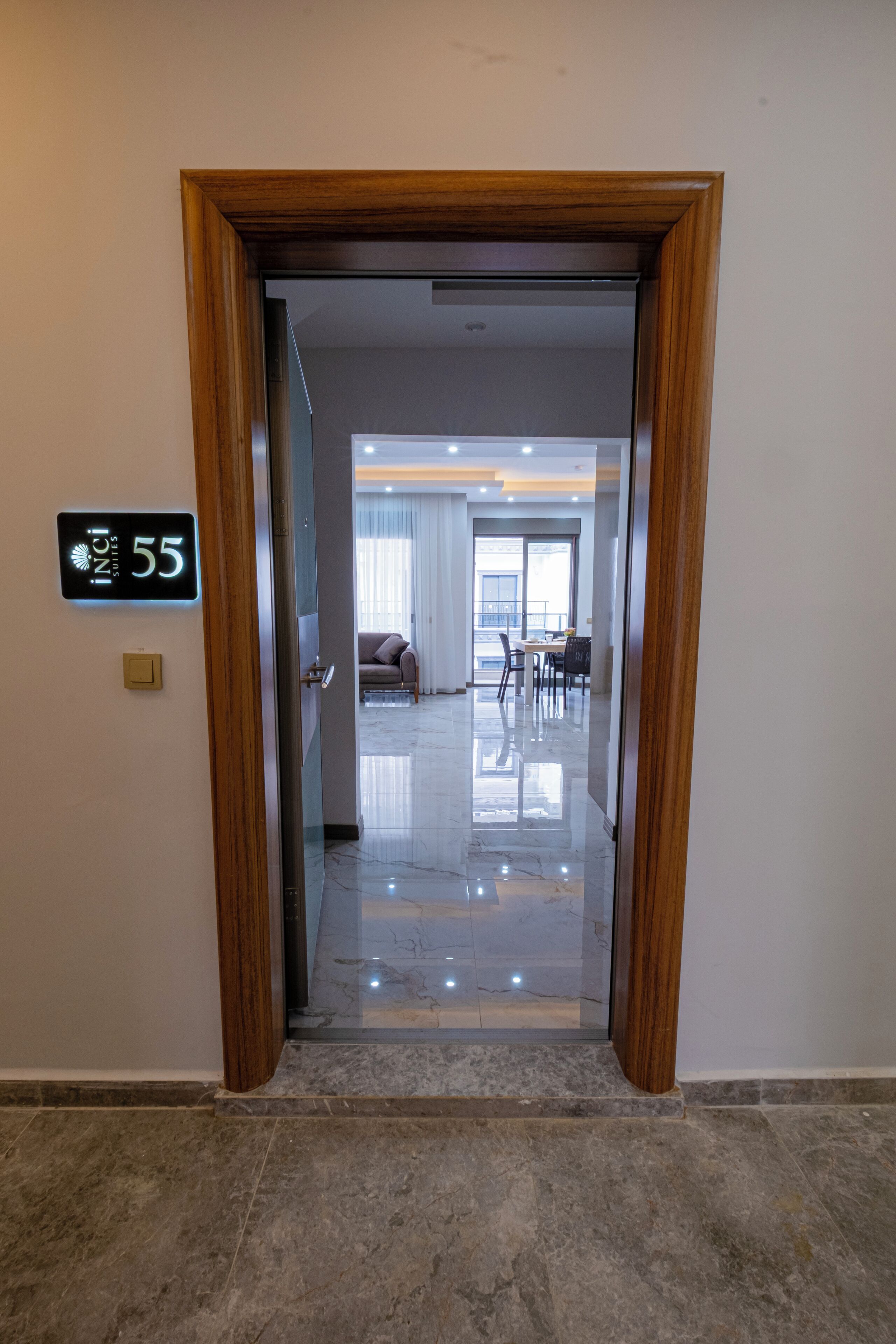 A hotel entrance with a wooden door, a digital display showing room number 55, and a view into a well-lit dining area with a sofa and chairs.