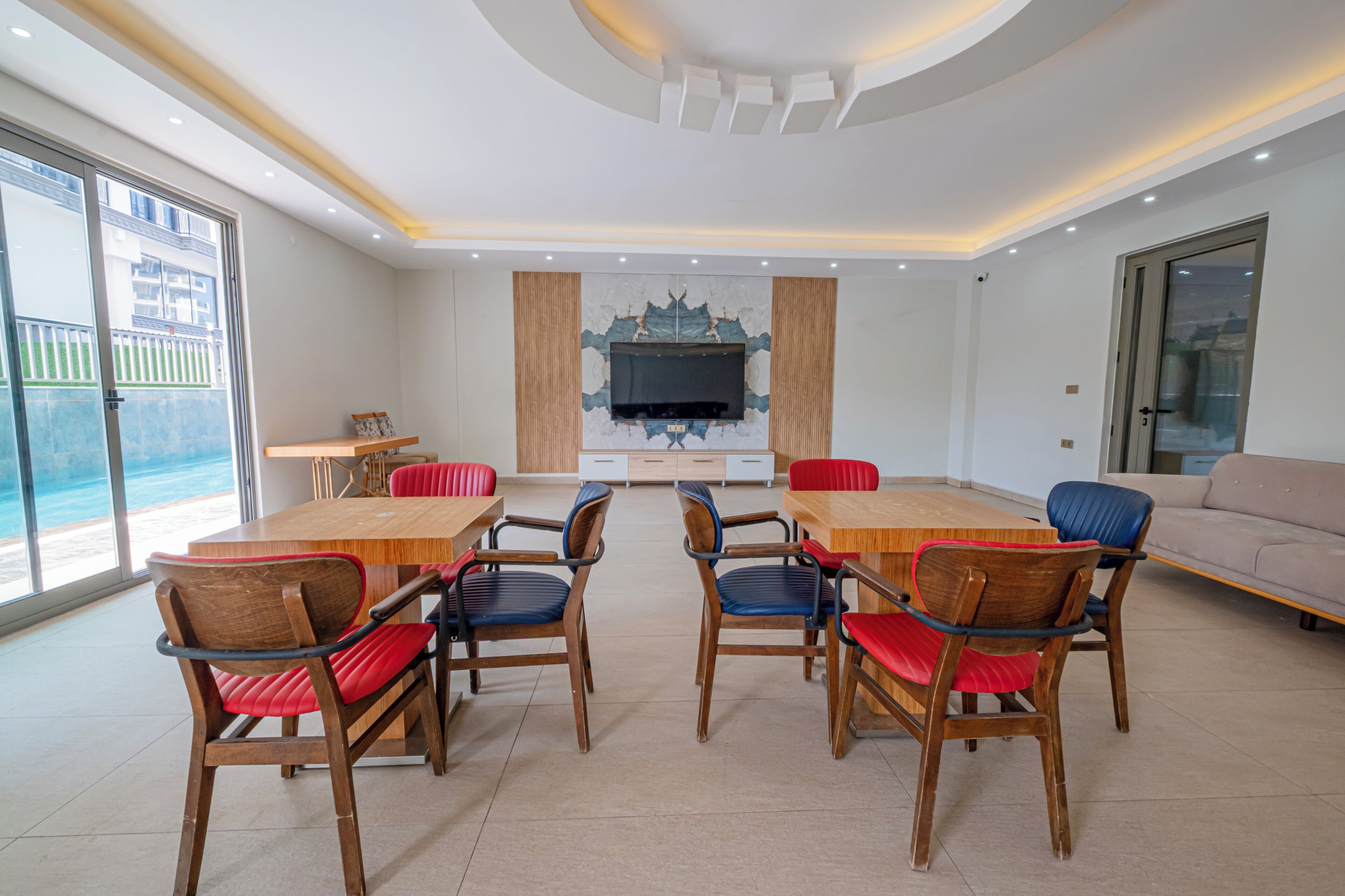 A spacious living room with a large wooden dining table surrounded by chairs, a flat-screen TV mounted on the wall, and a sofa.