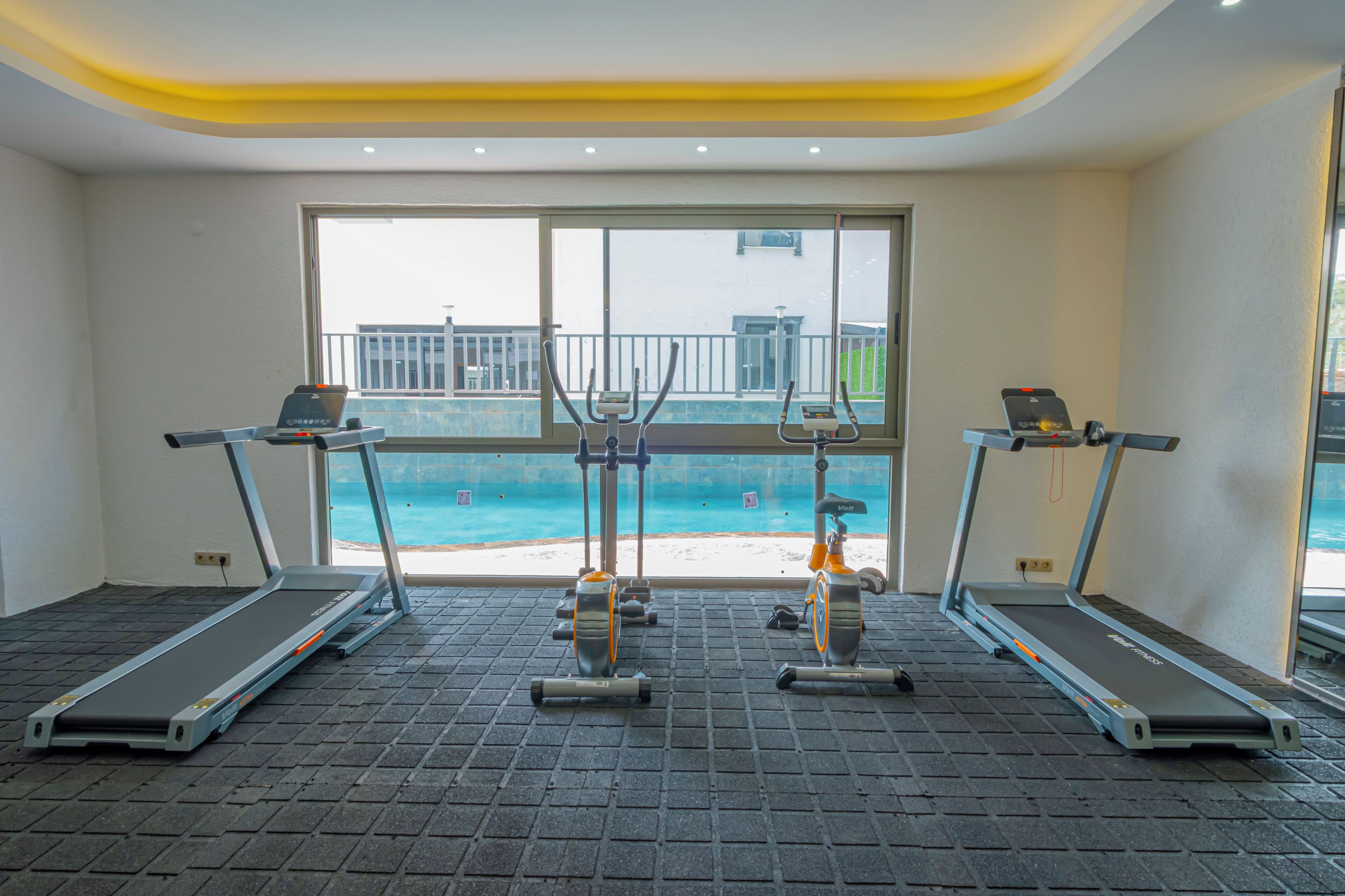 A gym with treadmills and exercise equipment, large windows with a view of the beach and buildings.
