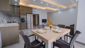 A modern kitchen with stainless steel appliances, a dining area with a wooden table and dark chairs, and a living area with a sofa and TV.