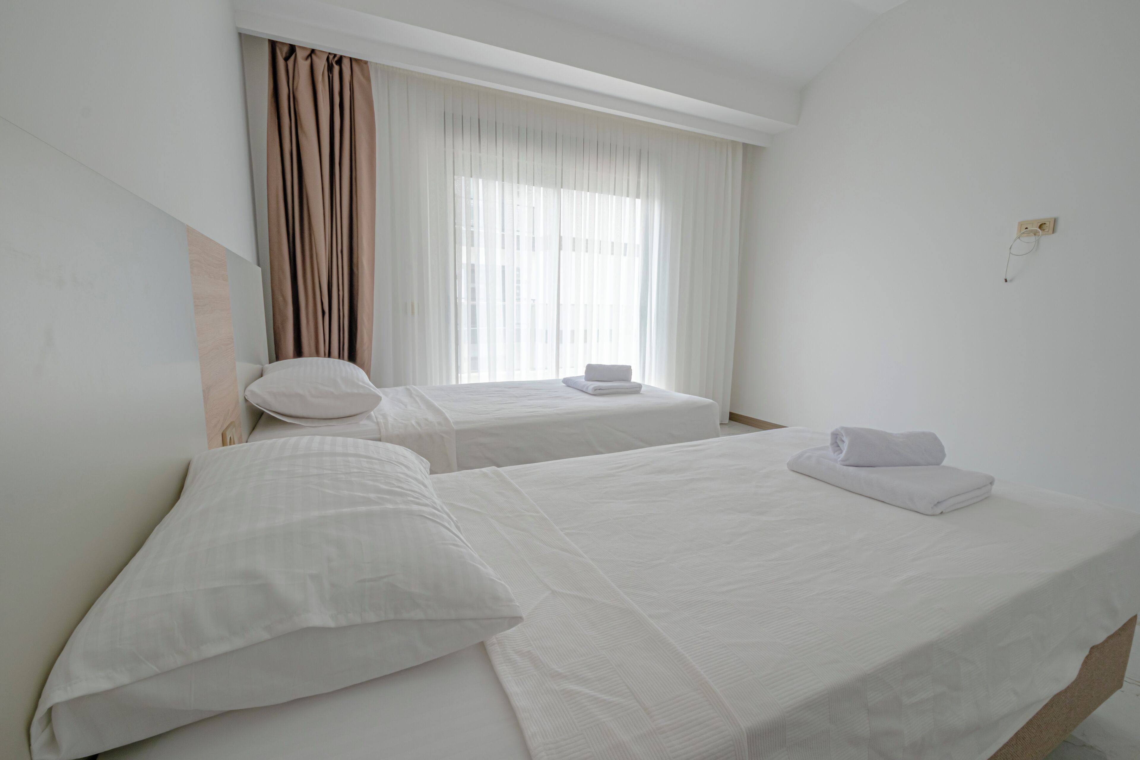 A hotel room with two single beds, a window with curtains, and a wall socket.