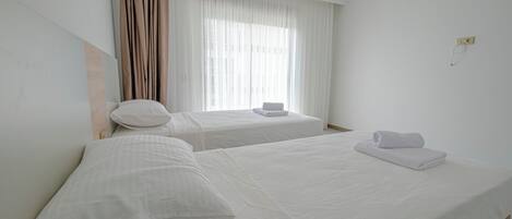 A hotel room with two single beds, a window with curtains, and a wall socket.
