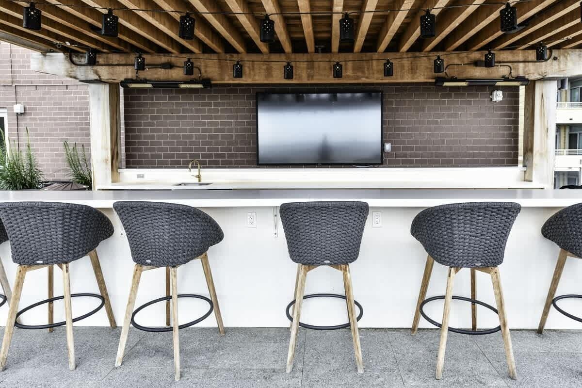 A bar area with a flat-screen TV mounted on a brick wall, wooden ceiling beams, and three black wicker bar stools.