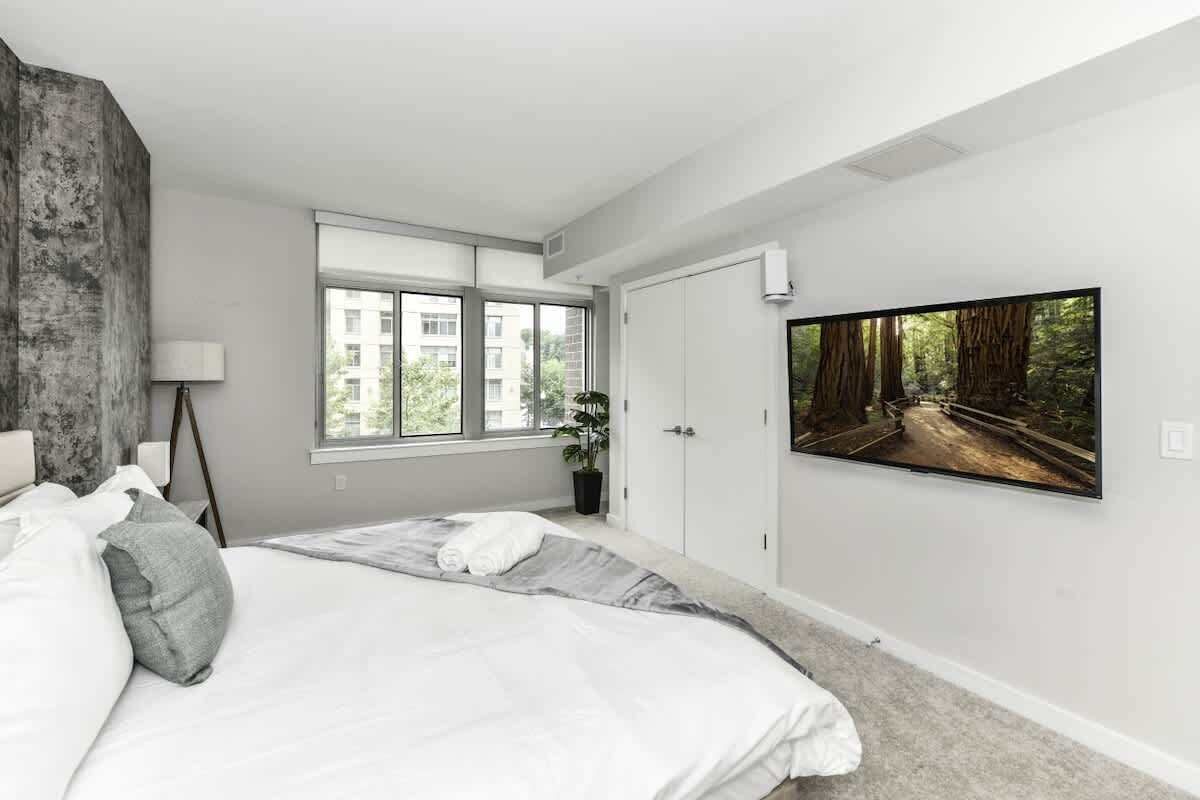 Une chambre moderne avec un grand lit, une fenêtre donnant sur des immeubles, un tableau représentant une forêt et une plante en pot.