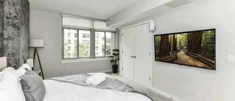 Une chambre moderne avec un grand lit, une fenêtre donnant sur des immeubles, un tableau représentant une forêt et une plante en pot.