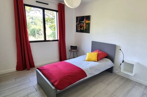 Une chambre à coucher avec un lit, des rideaux rouges, une fenêtre, un petit tabouret et une étagère fixée au mur.
