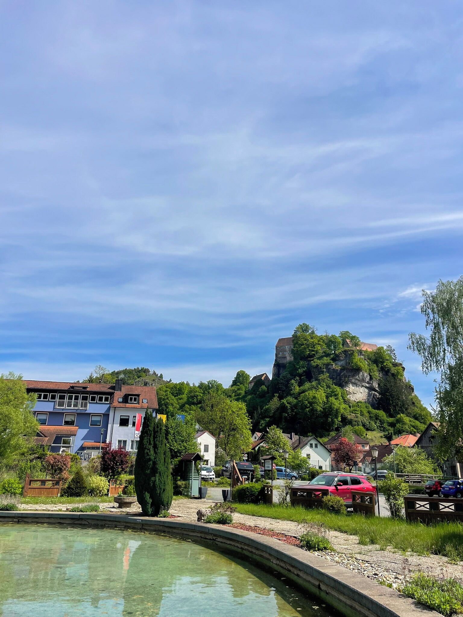 A residential area with a small pond, houses, and a hill with a castle-like structure in the background.