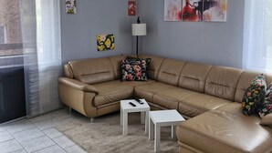 A living room with a beige sectional sofa, a small white side table, a floor lamp, and artwork on the walls. - Spacious 4-room apartment for the whole family with an eat-in kitchen (Elsdorf)