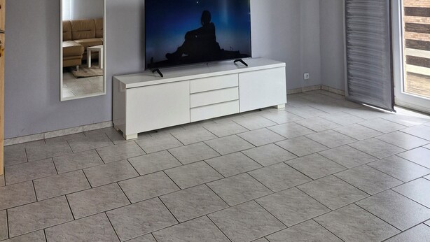 A modern living room with a large flat-screen TV on a white console, a mirror on the wall, and a large window with curtains. - Spacious 4-room apartment for the whole family with an eat-in kitchen (Elsdorf)