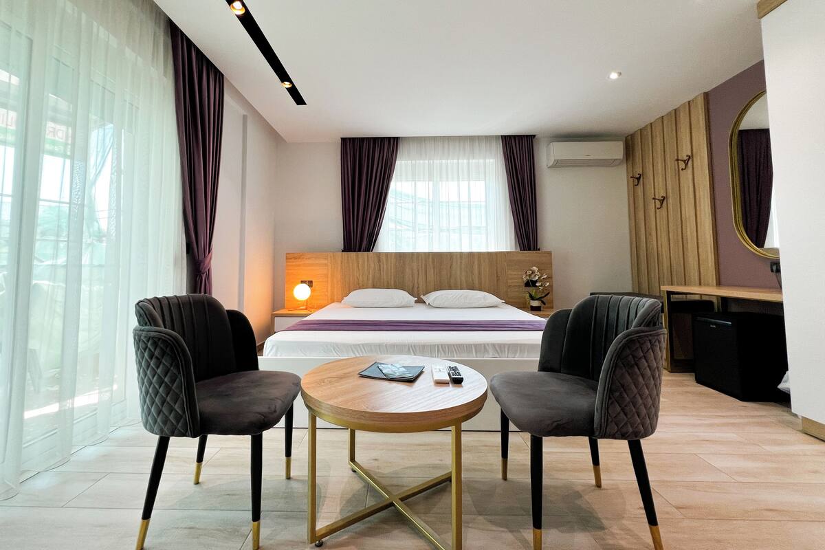 Een moderne hotelkamer met een bed, twee fauteuils, een klein rond tafeltje en een bureau.