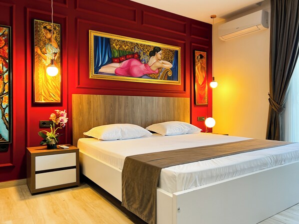 Quarto de hotel com cabeceira de madeira, cama com roupa de cama branca, mesa de cabeceira com um vaso de flores e duas luminárias fixadas na parede.
