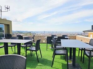 Área externa com mesas e cadeiras, vista para o horizonte urbano e um edifício com telhado plano.