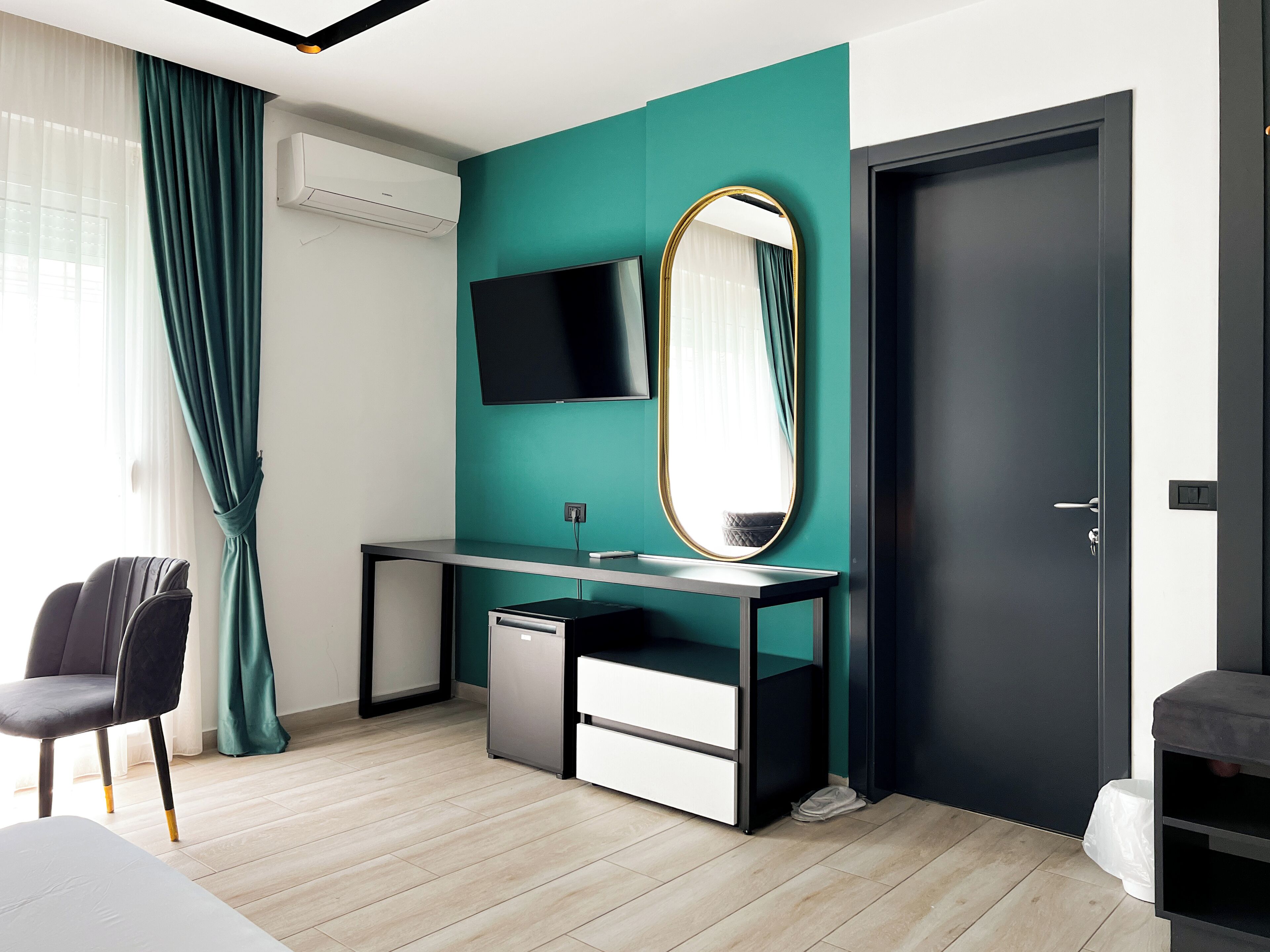 A modern hotel room with a teal accent wall, a large mirror, a flat-screen TV, a desk with a computer, and a chair.