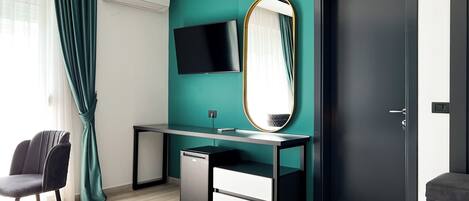 A modern hotel room with a teal accent wall, a large mirror, a flat-screen TV, a desk with a computer, and a chair.