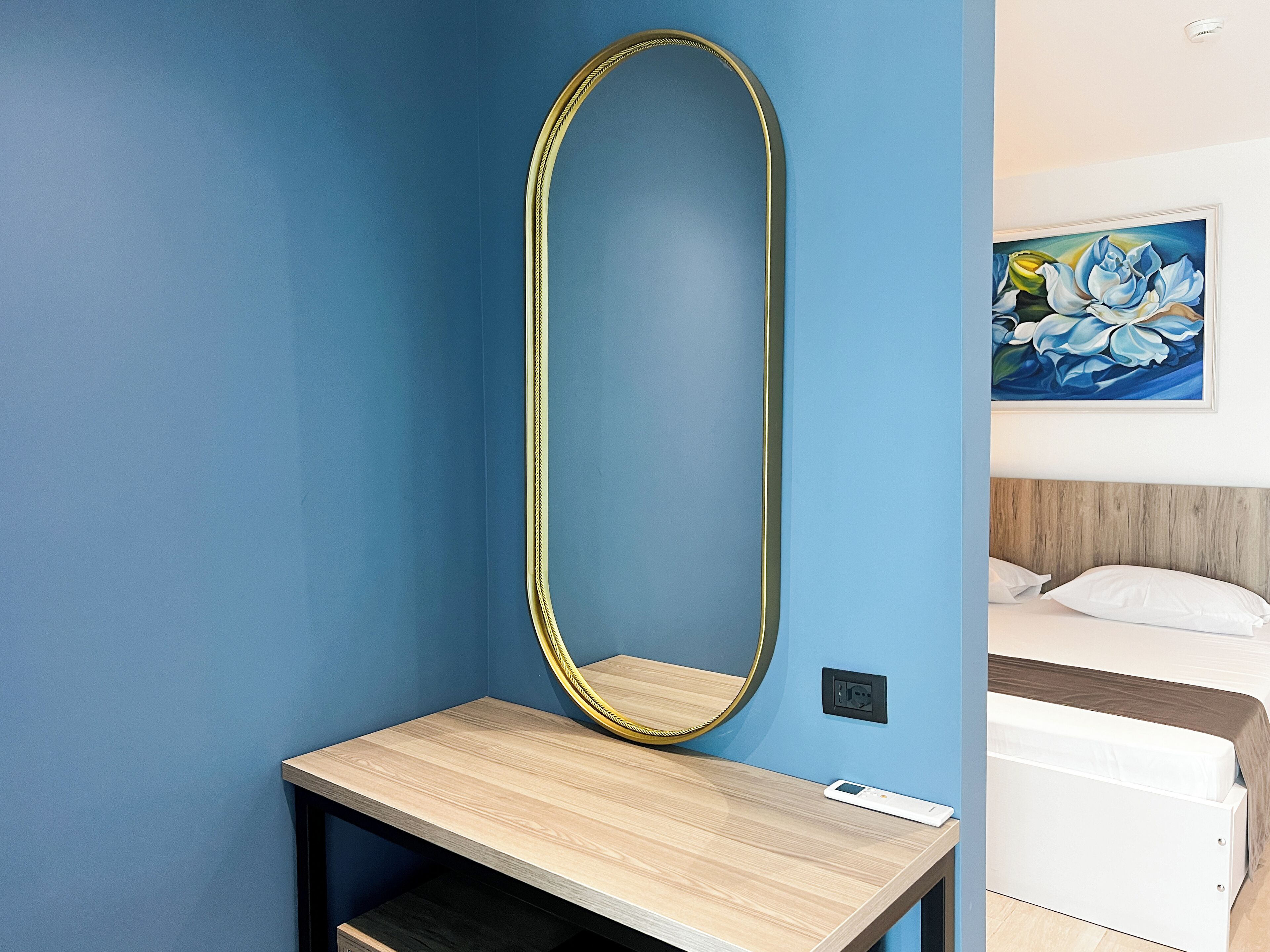 A hotel room with a large oval mirror, a wooden bedside table, a white bed, and a painting of flowers on the wall.
