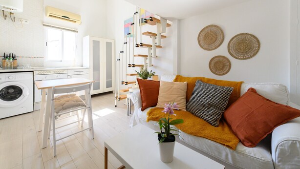 Una sala de estar moderna y bien iluminada con un sofá, una mesa de centro y una mesita con una planta en maceta. Se ve una cocina con lavadora al fondo.