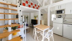 Cocina compacta con gabinetes blancos, microondas y lavavajillas. Hay una mesa de madera con sillas blancas y un televisor sobre un mueble.