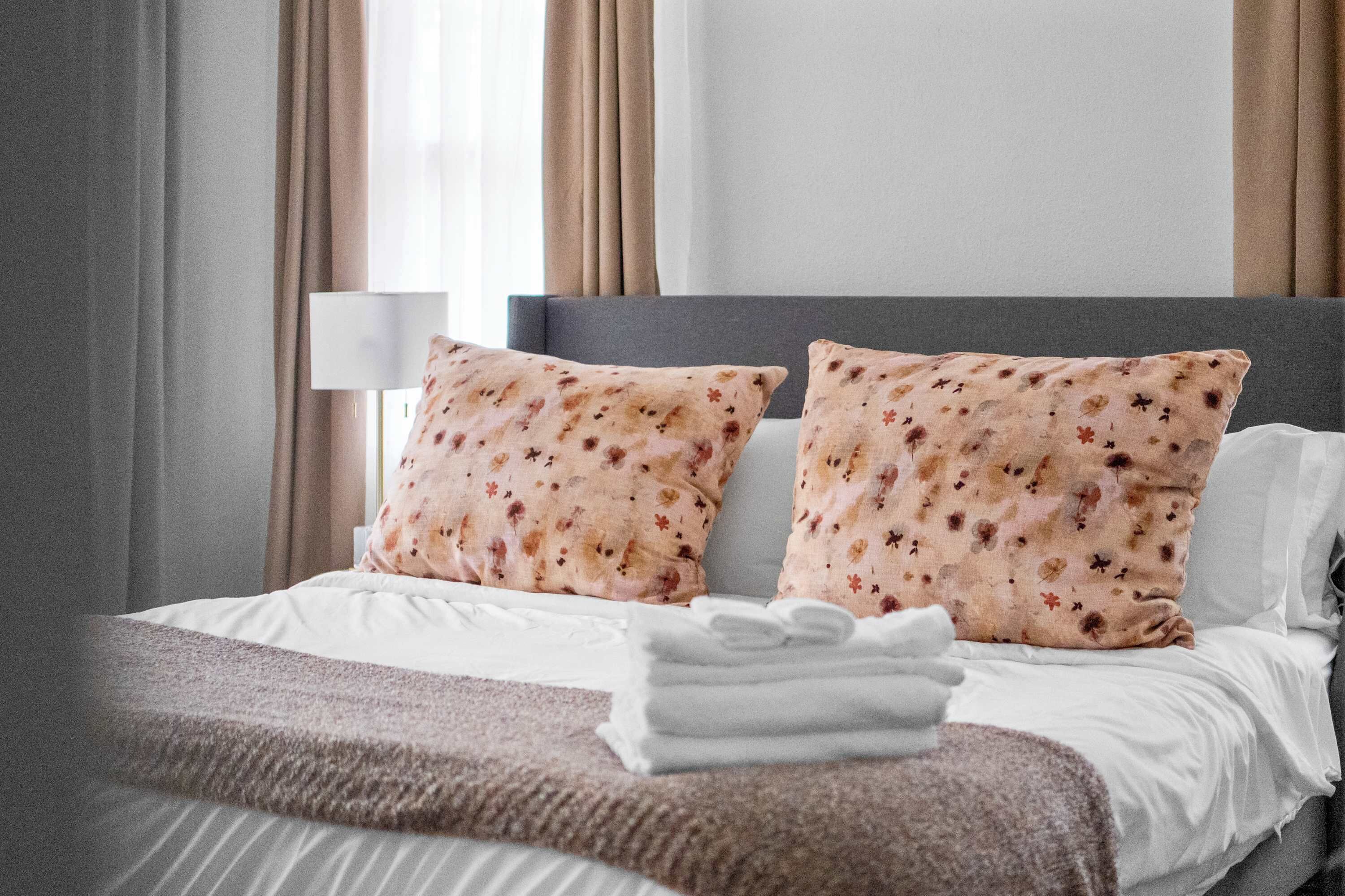 A neatly made bed with floral-patterned pillows, a folded white towel, and a grey headboard.