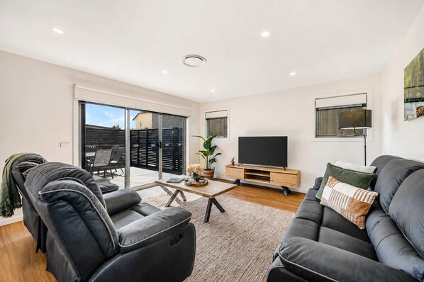 A modern living room with a sofa, a coffee table, and a television. There is a sliding glass door leading to an outdoor area.