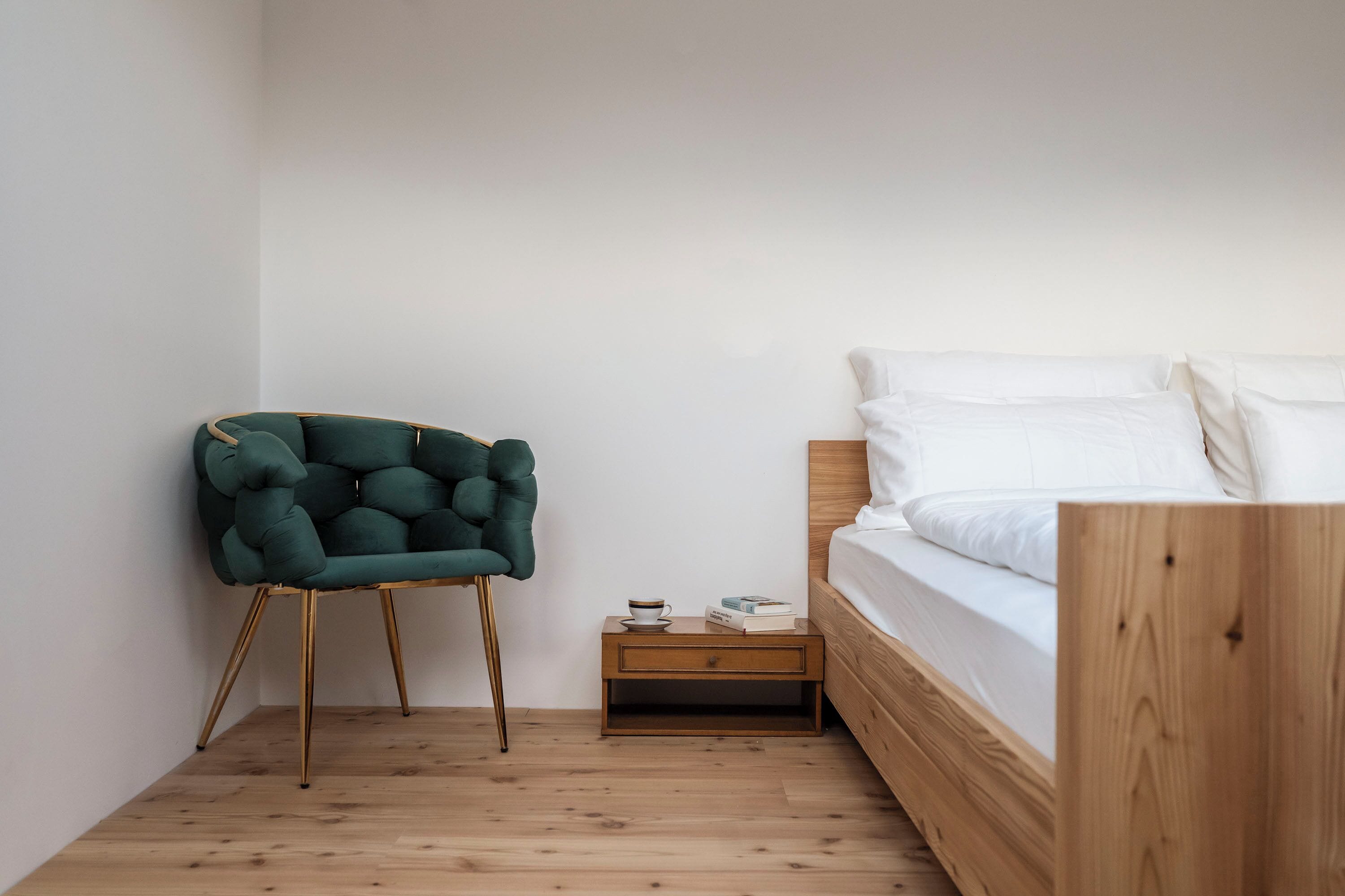 A bedroom with a wooden bed, a green upholstered chair, a nightstand with a cup and books, and a wooden floor.