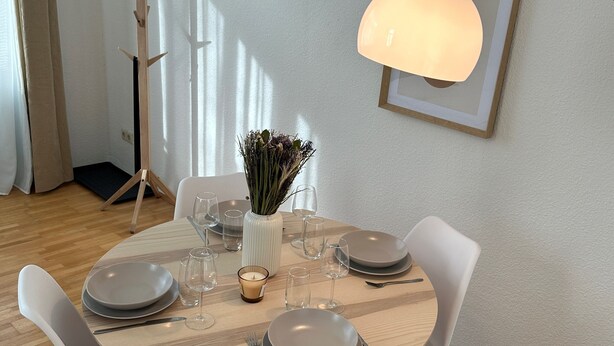 Een eethoek met een ronde tafel gedekt voor vier personen, witte stoelen, een vaas met gedroogde bloemen en een kaars.